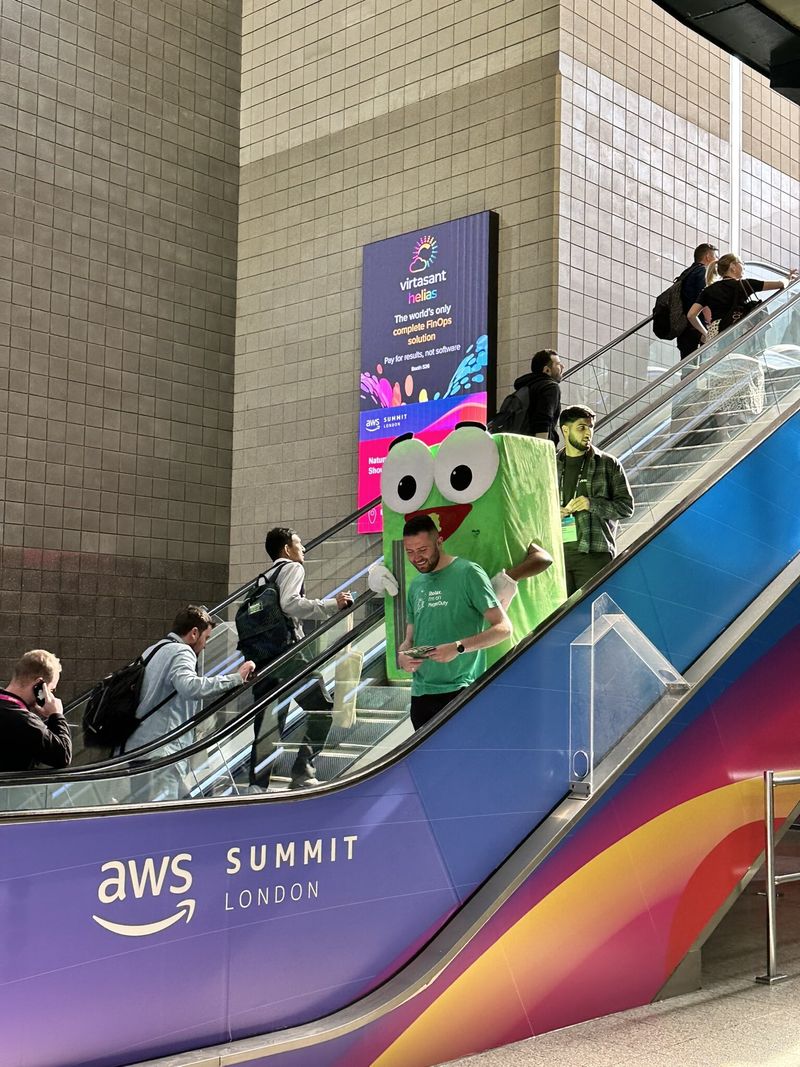Pagey, the PagerDuty mascot, rides an escalator at the London ExCel center, accompanied by a coworker in a PagerDuty t-shirt